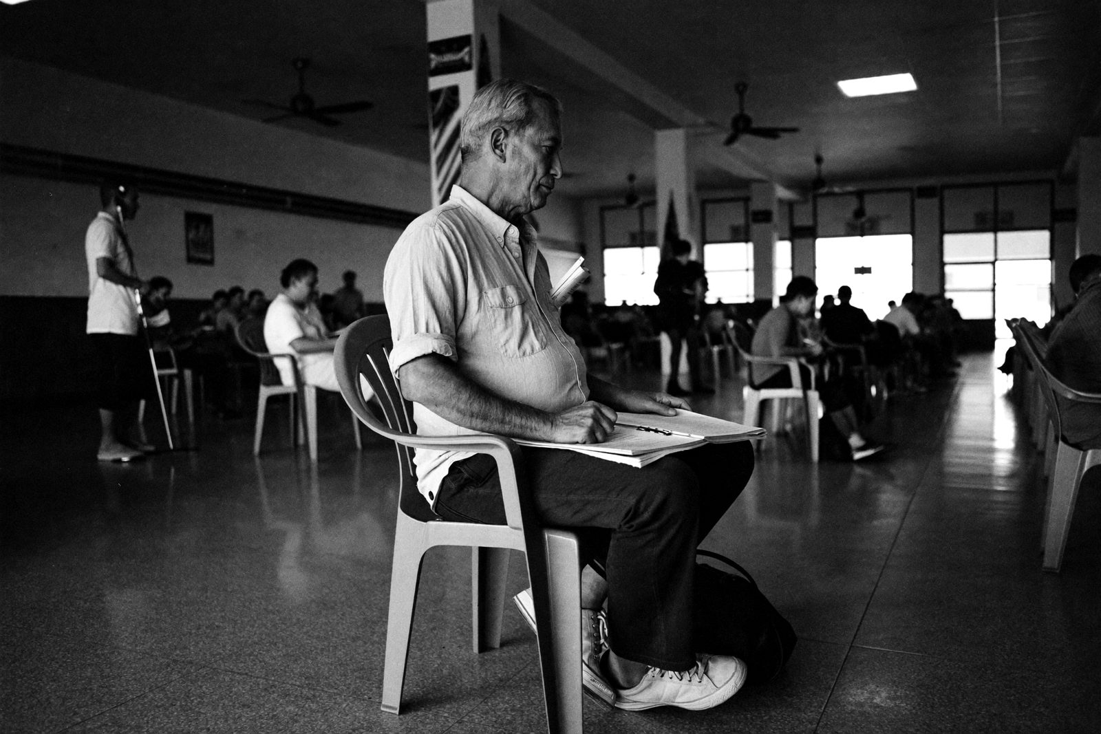 Ein älterer Mann im hellen Hemd wartet mit einer Dokumentenmappe auf einem Plastikstuhl in einer großen Behördenhalle in Bangkok, im Hintergrund weitere Wartende unter Deckenventilatoren.