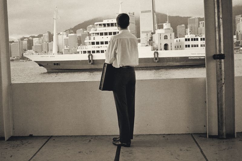 Ein Mann in Hemd und Hose steht mit einer Aktentasche an einem Fährpier in Hongkong, vor ihm eine Fähre und die Hochhäuser am Hafen im Dunst.