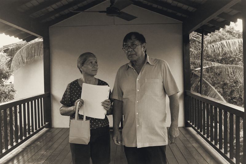 Ein älteres Paar steht mit einer Mappe und einer kleinen Papiertüte auf einer Holzveranda in Cebu, ein Deckenventilator über ihnen, draußen Regen auf den Blättern.
