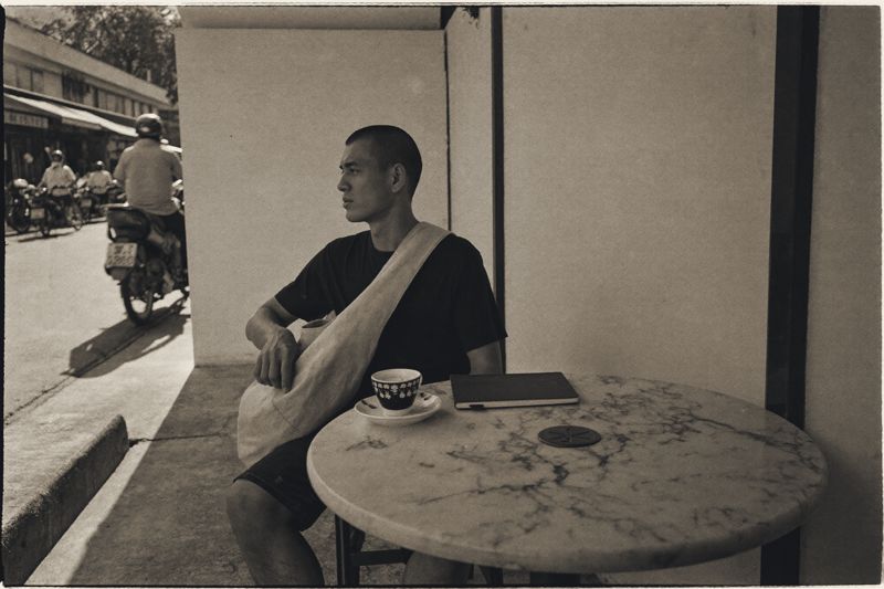 Ein junger Mann mit Umhängetasche sitzt am Morgen an einem Marmortisch eines Straßencafés in Chiang Mai, vor ihm ein geschlossenes Notizbuch und eine Kaffeetasse, dahinter fahren Motorräder vorbei.