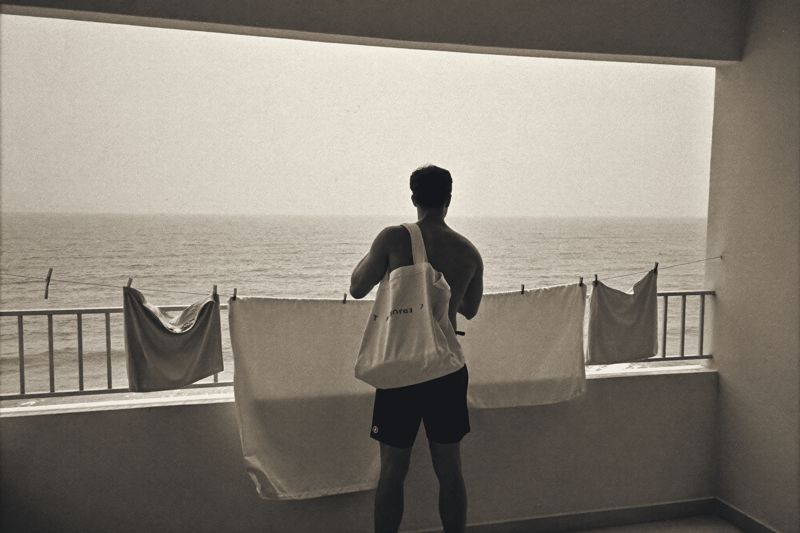 Ein junger Mann mit Stofftasche steht auf einem Wohnungsbalkon in Da Nang, Wäsche auf der Leine, dahinter das Meer im Dunst.