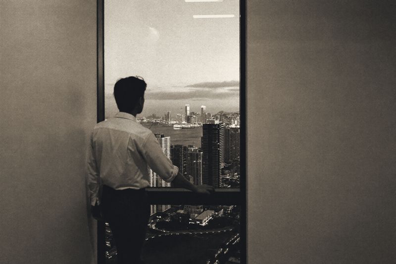 Ein Mann in hellem Hemd steht in der Dämmerung an einem hohen Fenster in einem Büroturm in Hongkong, eine Hand auf der Fensterbrüstung, unter ihm der Hafen und die Hochhäuser im Dunst.