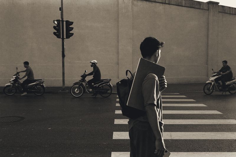 Ein junger Mann mit Umhängetasche wartet am späten Nachmittag an einer Kreuzung in Ho-Chi-Minh-Stadt, vor ihm Motorräder an einer roten Ampel, der Asphalt nass vom Regen.