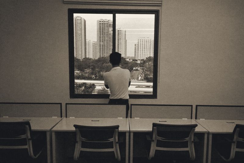 Ein Mann im hellen Poloshirt steht von hinten gesehen am Fenster eines leeren Großraumbüros in Makati, vor ihm Hochhäuser und Bäume, im Vordergrund eine Reihe leerer Schreibtische mit Stühlen.