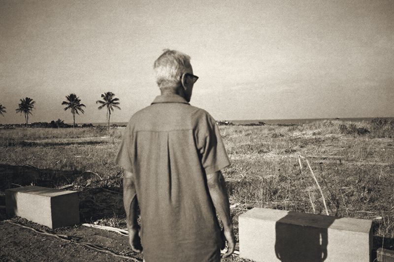 Ein älterer Mann mit grauem Haar und Sonnenbrille steht von hinten gesehen am Rand eines brachliegenden Grundstücks bei Hua Hin, vor ihm trockenes Gras, Kokospalmen und in der Ferne das Meer, neben ihm zwei niedrige Betonblöcke.