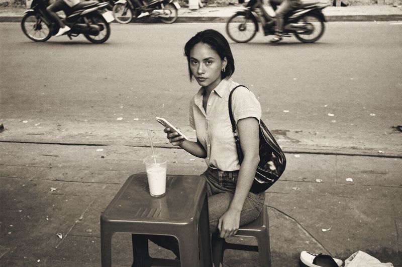 Eine junge Frau mit kurzem Haar sitzt auf einem niedrigen Plastikhocker an einem Straßencafé in Hanoi, in der Hand ein Mobiltelefon, vor ihr ein Glas Eiskaffee, hinter ihr fahren Motorräder vorbei.