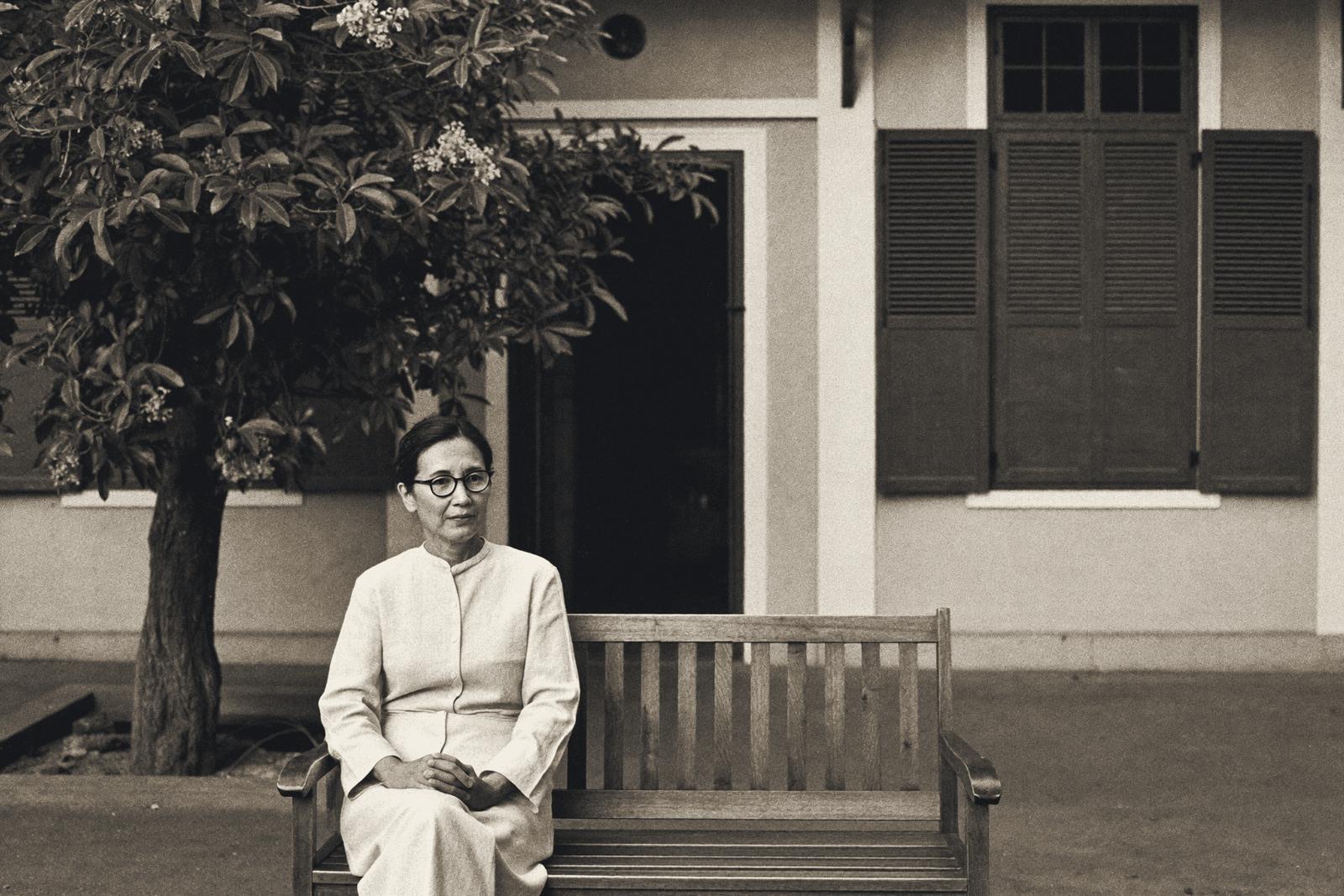 Eine Frau mit Brille und hellem Leinenkleid sitzt allein auf einer Holzbank im Hof eines alten Kolonialgebäudes in Hanoi, die Hände im Schoß gefaltet, hinter ihr ein blühender Frangipani-Baum und hohe Holzläden.