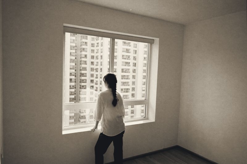 Eine Frau mit langem Zopf steht von hinten gesehen am Fenster einer leeren Wohnung mit weißen Wänden und Holzboden und schaut auf dicht stehende Wohnhochhäuser.
