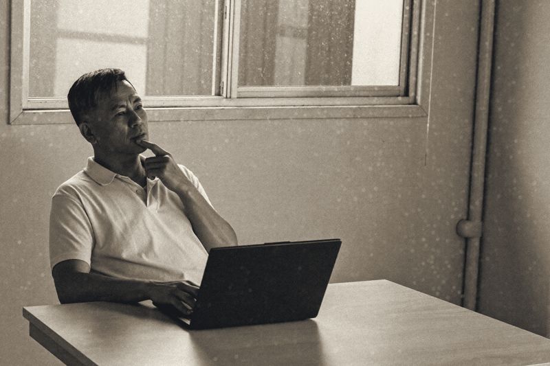 Ein Mann im weißen Poloshirt sitzt an einem hellen Tisch vor einem aufgeklappten Laptop, eine Hand am Kinn, hinter ihm ein Fenster mit Regentropfen und ein kahler Raum.