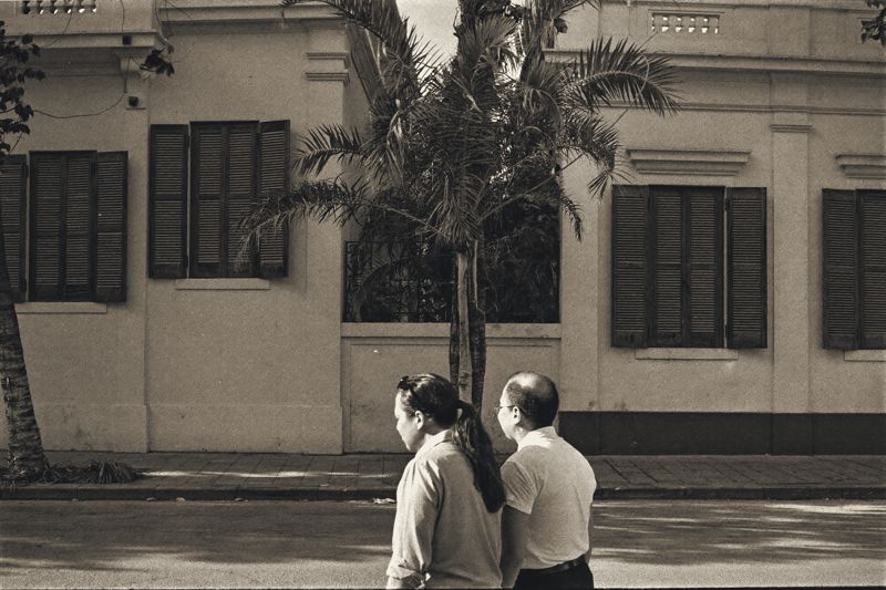 Eine Frau mit Pferdeschwanz und ein Mann mit Brille gehen von hinten gesehen eine ruhige Straße entlang, vor ihnen eine helle Kolonialvilla mit dunklen Holzläden und einer Palme.