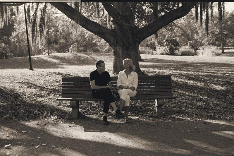 Ein Mann in dunklem Polohemd und eine Frau in weißer Bluse sitzen auf einer Parkbank unter einem großen Baum und unterhalten sich, auf dem Weg Licht und Schatten.