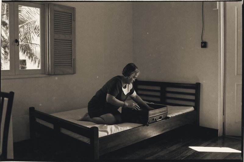 Eine ältere Frau mit kurzem Haar kniet auf einem Holzbett in einem schlichten Zimmer und packt einen Koffer, links ein Fenster mit halb geöffneten Läden und einer Palme, auf dem Boden ein Streifen Nachmittagslicht.