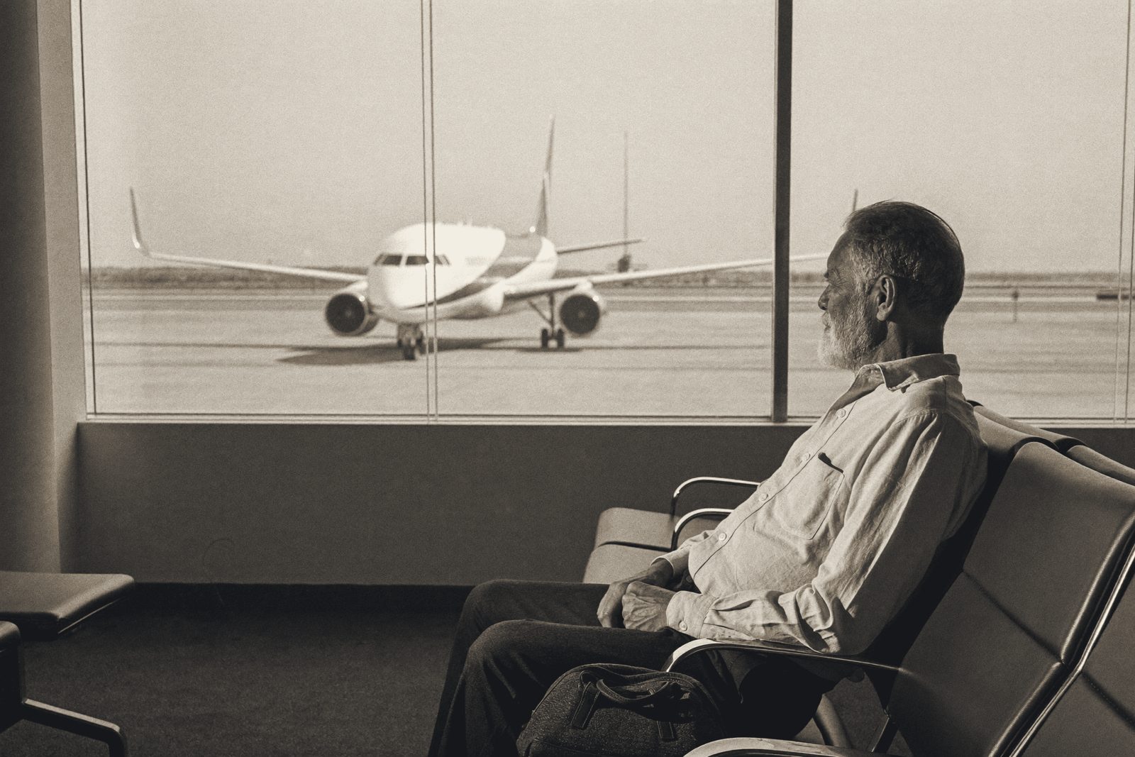Ein älterer Mann mit grauem Bart und hellem Hemd sitzt allein in einer Reihe von Wartesitzen am Flughafen, neben ihm eine Reisetasche, und schaut durch eine große Glasfront auf ein Flugzeug auf dem Vorfeld.
