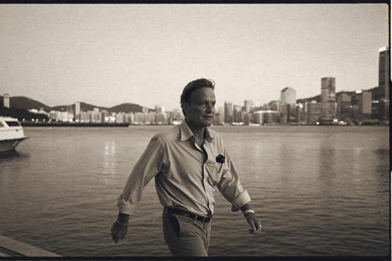 Ein Mann in hellem Hemd geht am Abend die Hafenpromenade entlang, hinter ihm das Wasser, ein Boot und die Hochhäuser am anderen Ufer.
