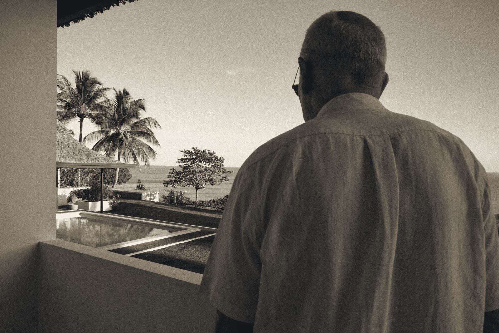 Ein älterer Mann mit Brille und hellem Leinenhemd steht von hinten gesehen auf der überdachten Terrasse einer Villa und schaut über Pool, Palmen und Garten aufs Meer.