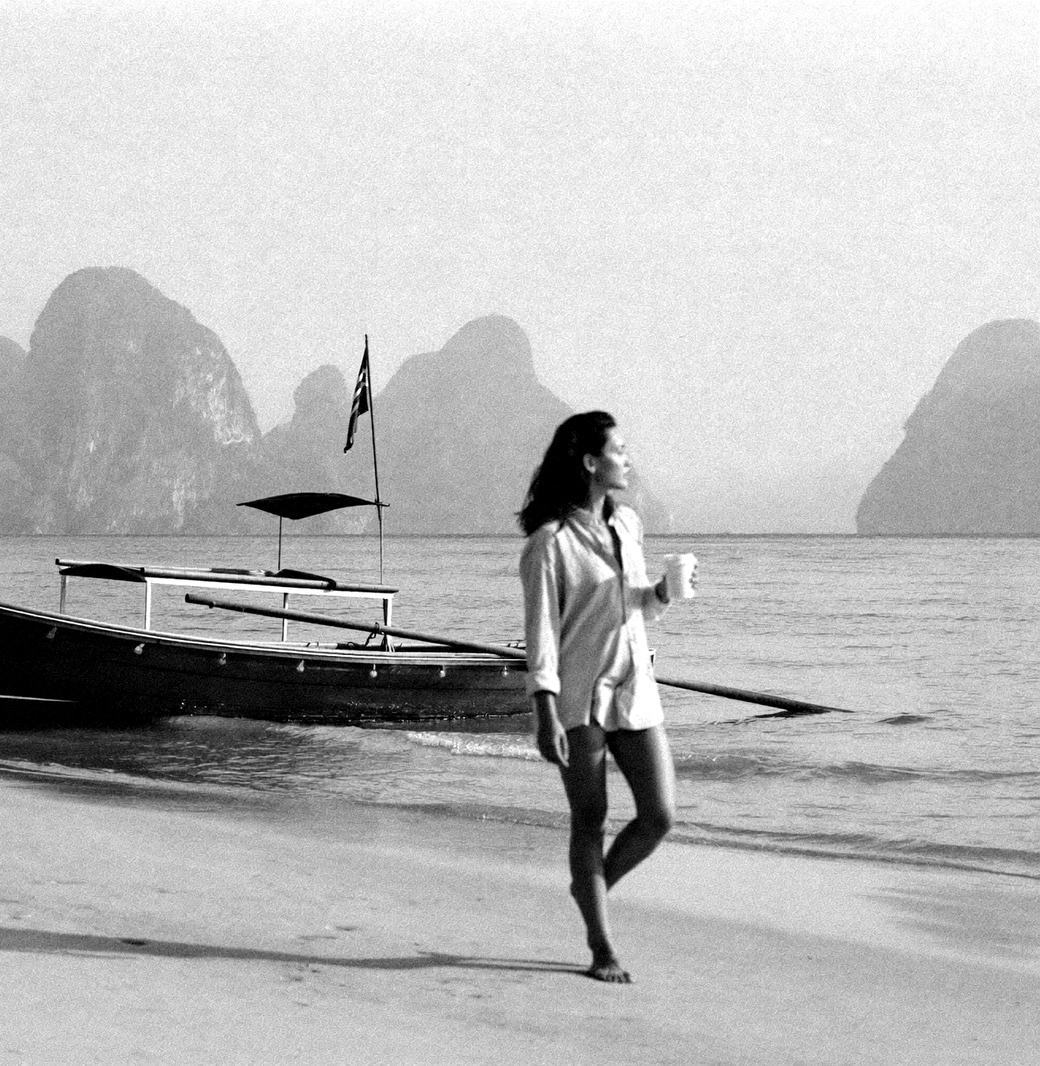 Früher Morgen an einem ruhigen Strand in Südthailand: ein Longtail-Boot am Wasser, eine Frau geht mit einer Kaffeetasse am Ufer entlang, Kalksteinfelsen im Dunst