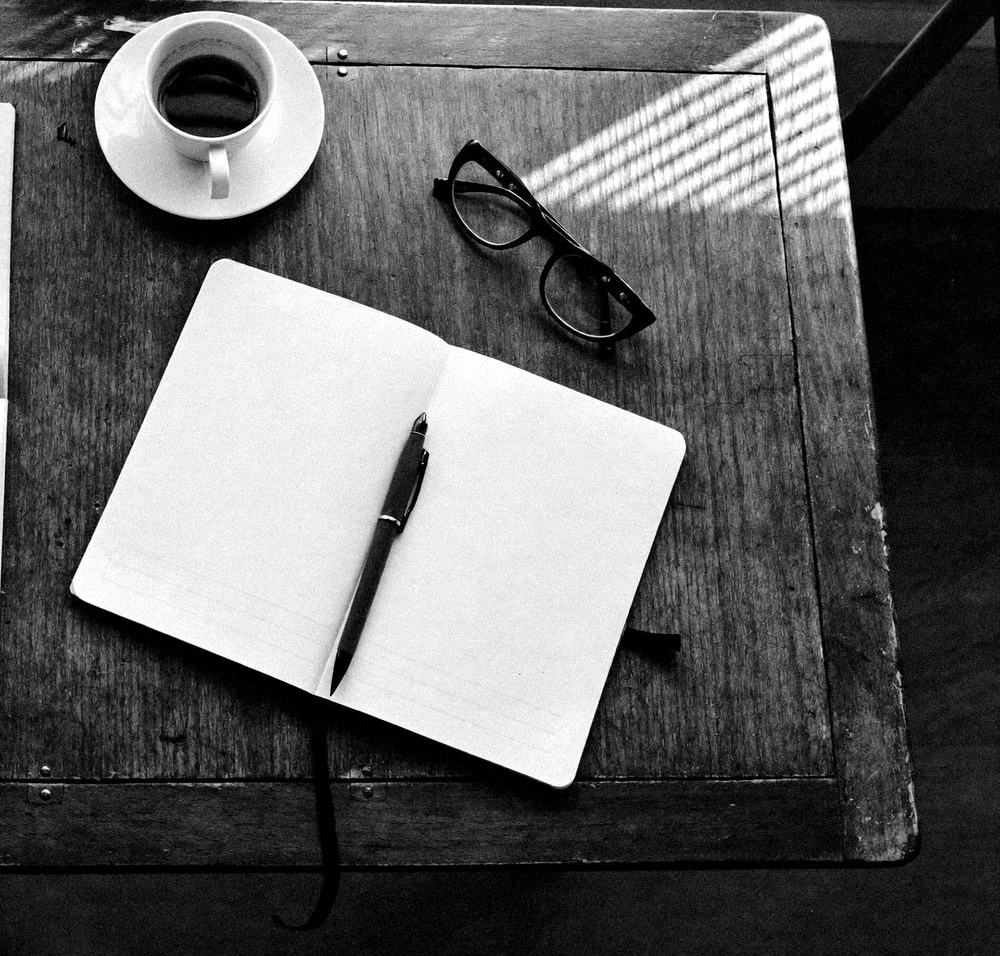 Schreibtisch in einer tropischen Wohnung: aufgeschlagenes Notizbuch, Füller, Lesebrille und Kaffeetasse im Morgenlicht