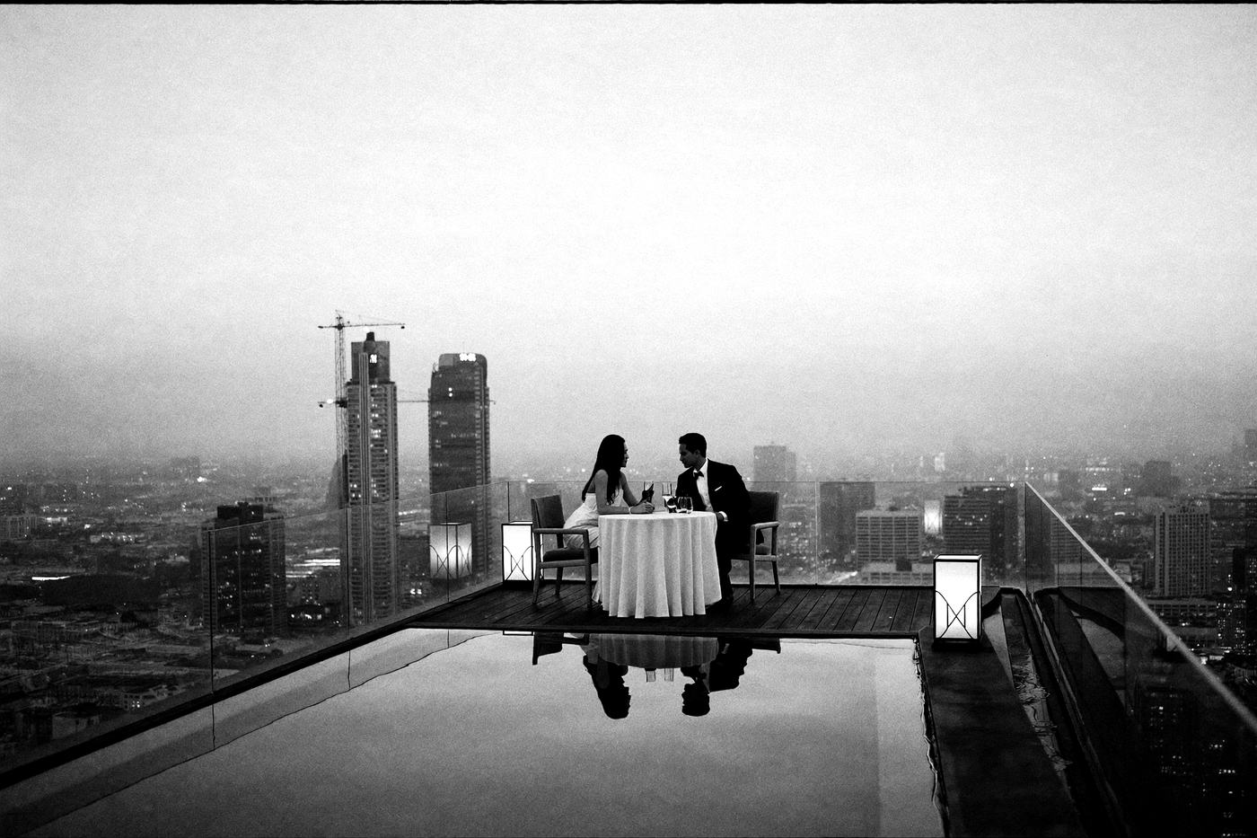 Abenddämmerung auf einer Dachterrasse hoch über Bangkok: Infinity-Pool, ein Paar am Tisch, die Skyline im Dunst